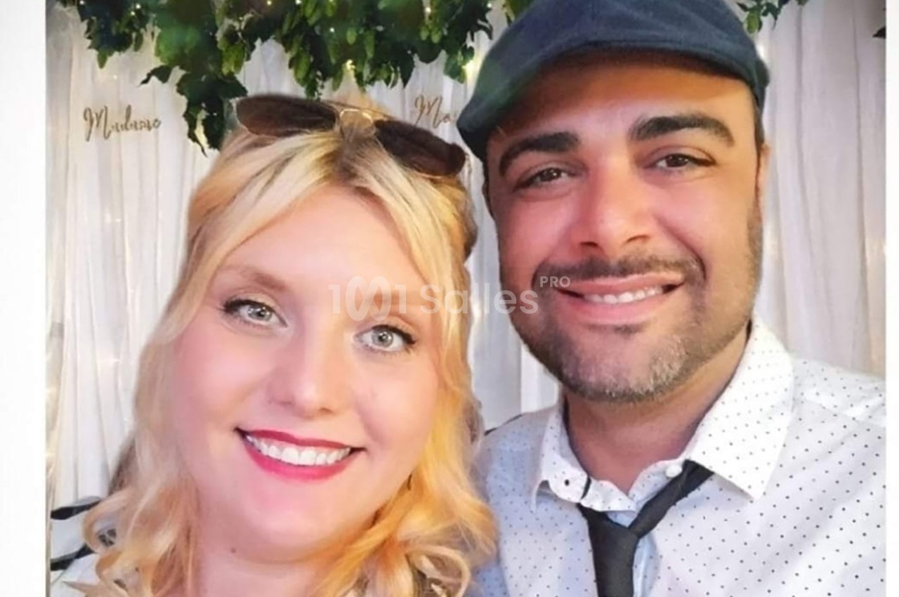 Un homme et une femme sourient devant un fond décoré de feuillage et de rideaux blancs.