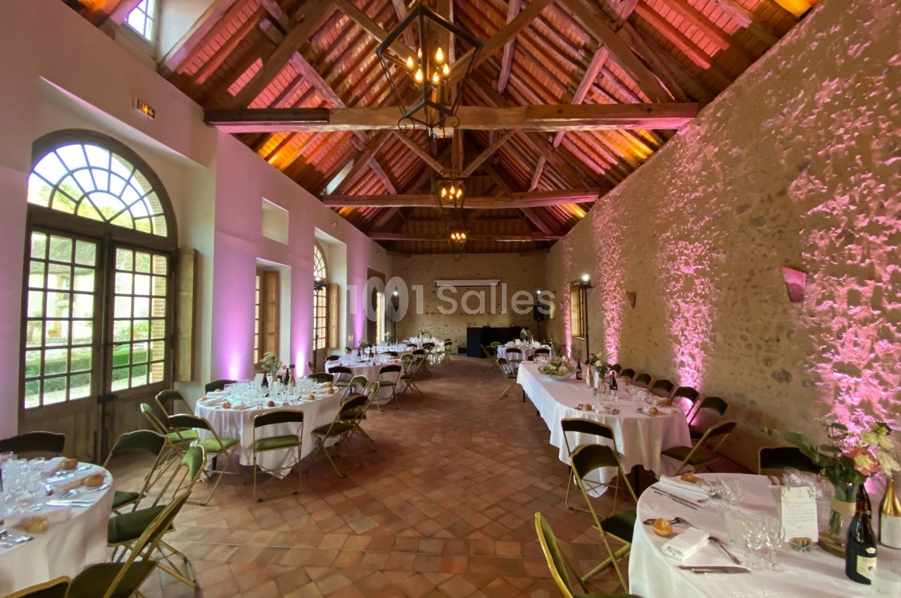 Salle de réception avec tables rondes dressées, chaises pliantes et éclairage chaleureux sous une charpente en bois.