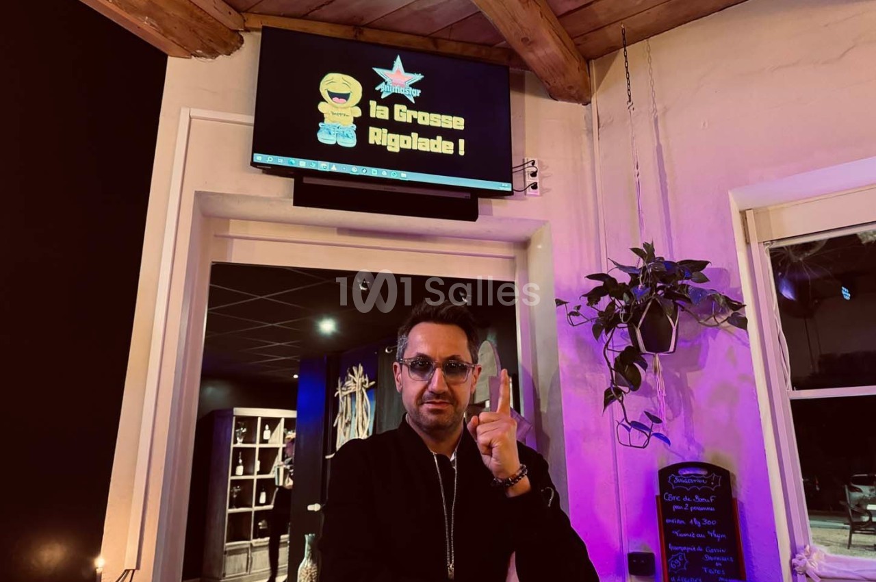 Un homme pose devant un écran affichant ’La Grosse Rigolade !’ dans un intérieur avec plantes et ardoise décorative.