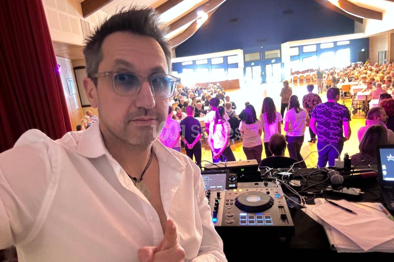 Un homme en chemise blanche devant une table de mixage, face à un public dans une salle lumineuse.