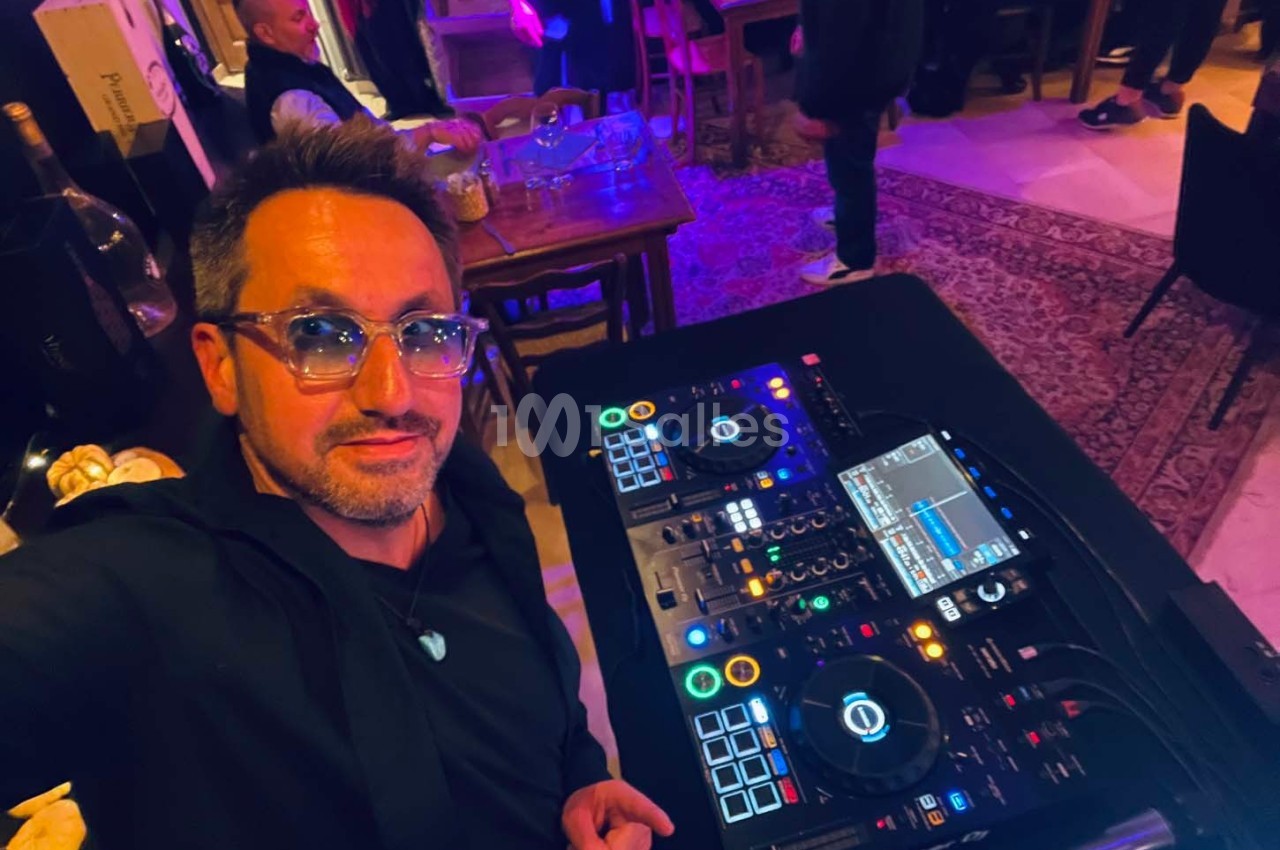 Un homme pose devant une table de mixage DJ dans un environnement intérieur avec éclairage tamisé.