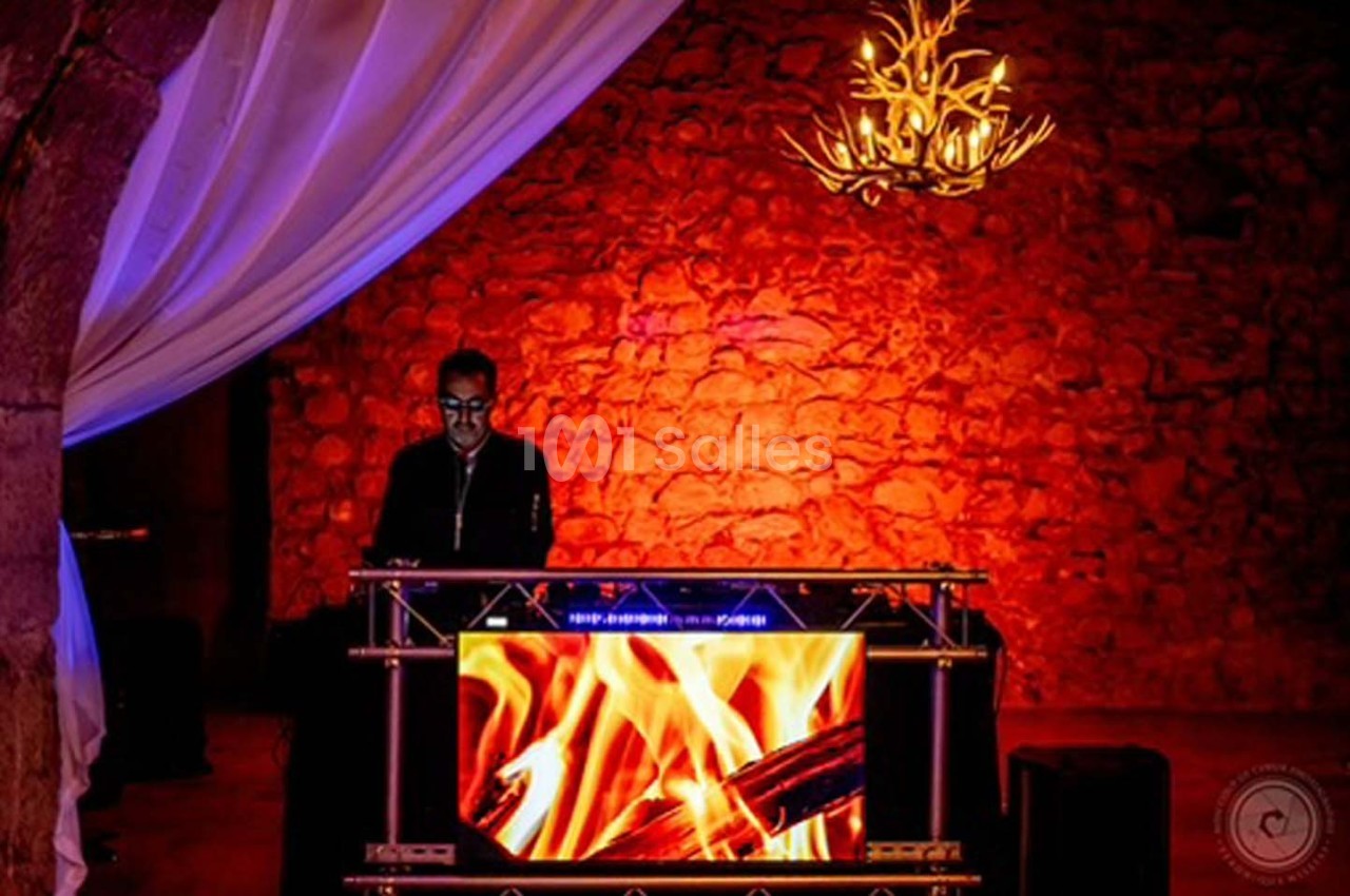 Un DJ mixe derrière une table ornée d'un écran affichant des flammes, dans une salle aux murs en pierre éclairée en rouge.
