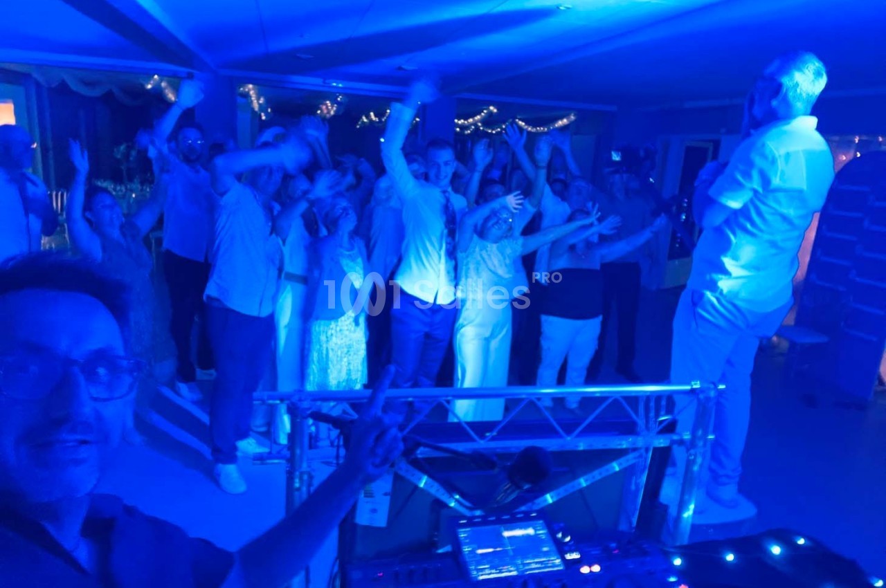 Groupe de personnes dansant sous des lumières bleues lors d'une fête, avec un DJ au premier plan.