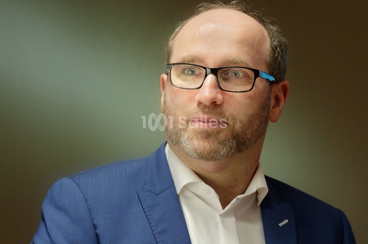 Homme portant des lunettes et un costume bleu, regardant légèrement sur le côté dans un environnement intérieur.