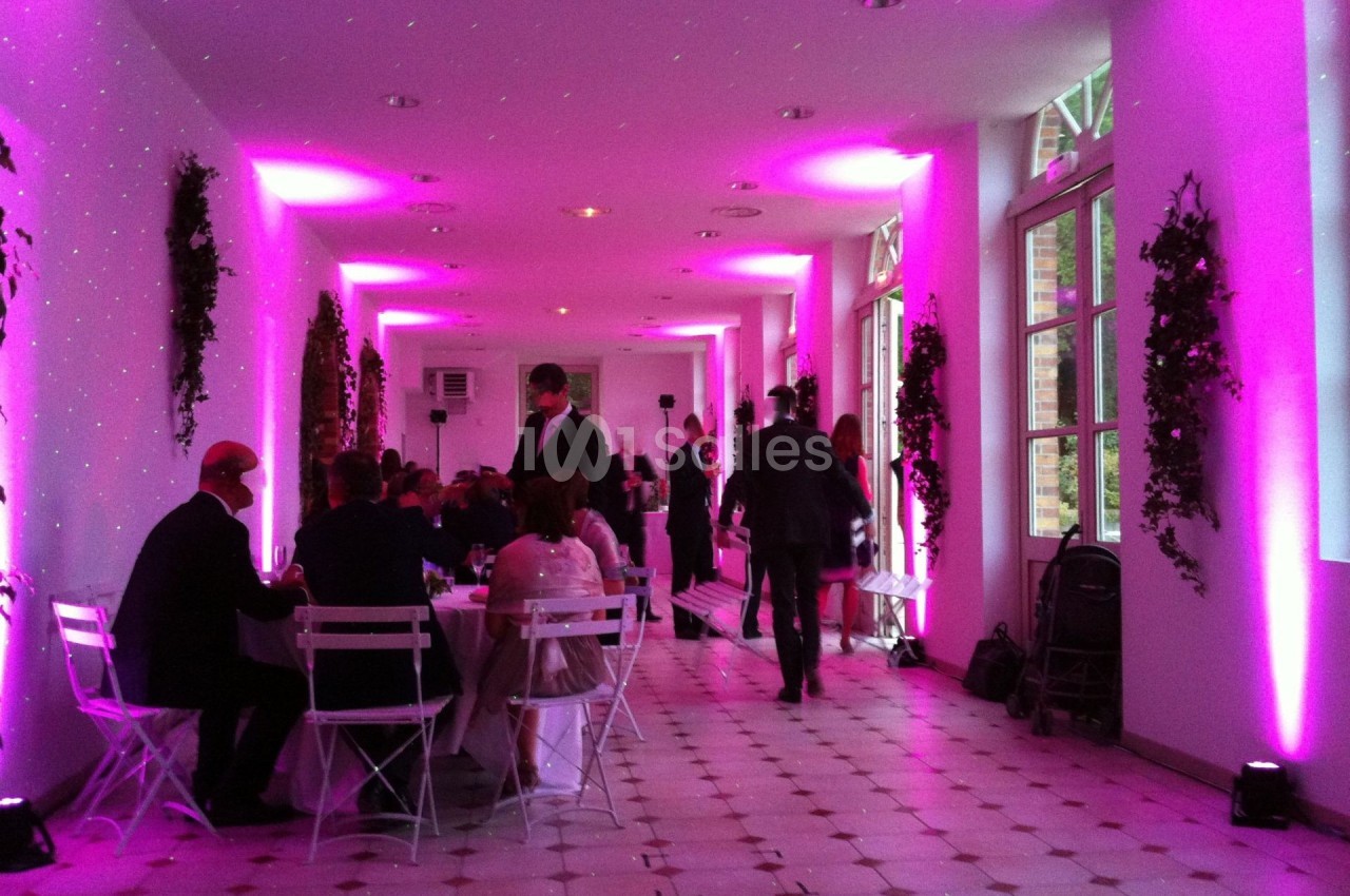 Salle éclairée de lumières roses, avec des invités assis à des tables blanches et d'autres debout dans un couloir.