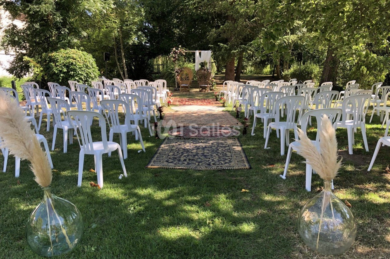 Chaises blanches disposées en extérieur, formant une allée centrale décorée de tapis et de vases avec des herbes séchées.