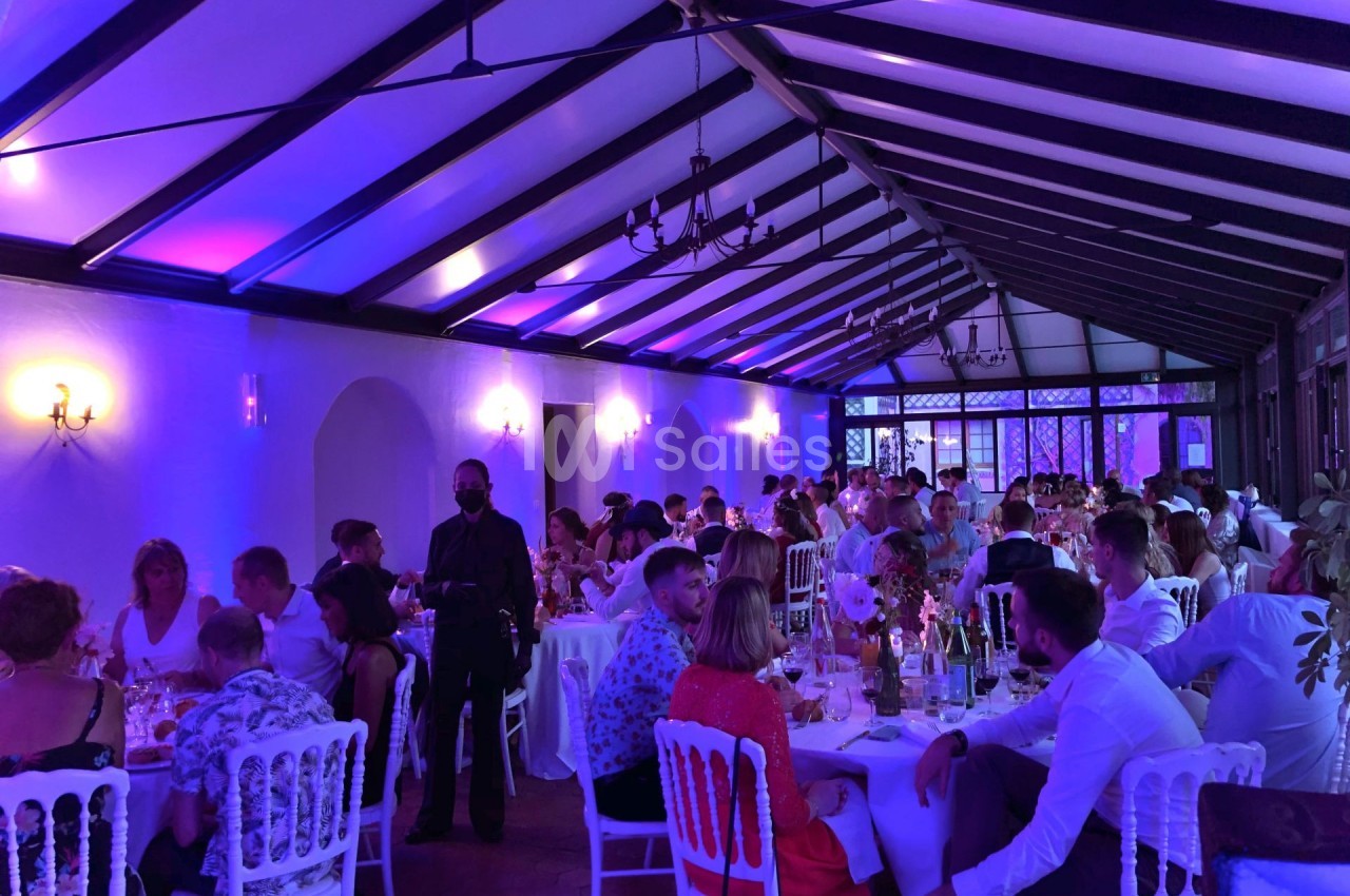 Salle de réception éclairée en violet, remplie de convives assis à des tables rondes lors d'un événement festif.