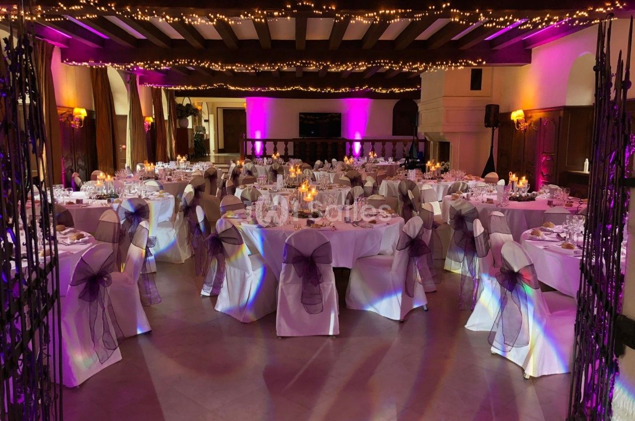 Salle de réception décorée pour un événement, avec tables dressées, chaises ornées de rubans et guirlandes lumineuses au…