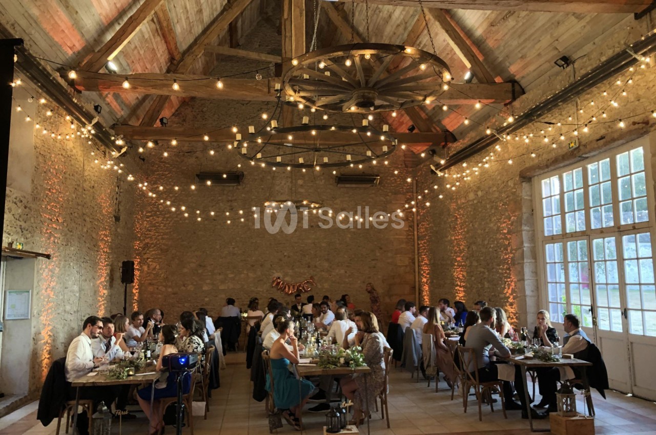 Salle de réception en pierre avec des invités dînant à des tables décorées, éclairée par des guirlandes lumineuses.