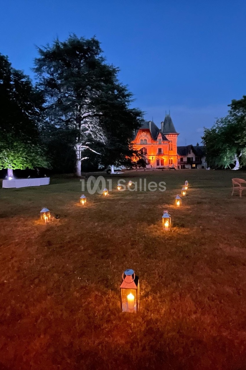 Allée de lanternes éclairées menant à un château illuminé en orange, entouré d'arbres au crépuscule.