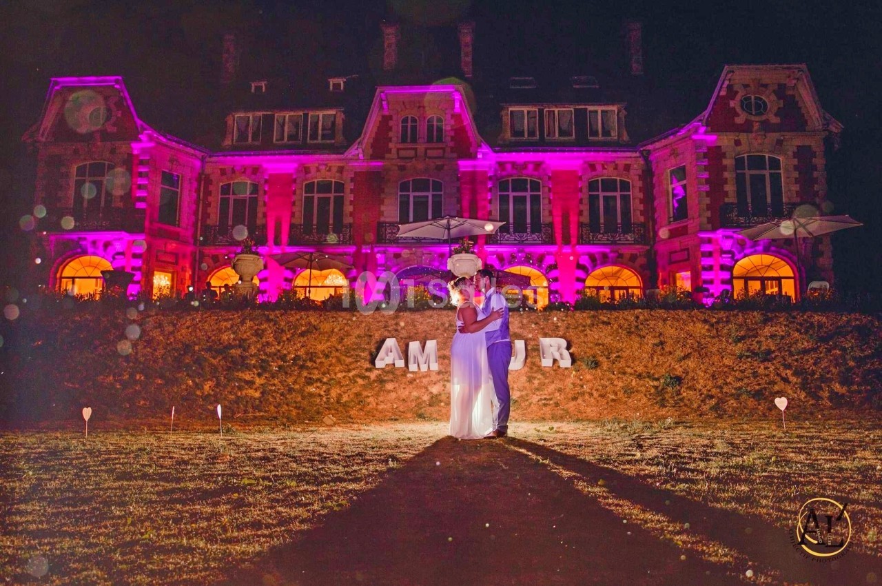 Un couple s'embrasse de nuit devant un bâtiment illuminé en rose, avec le mot ’AMOUR’ en lettres blanches au sol.