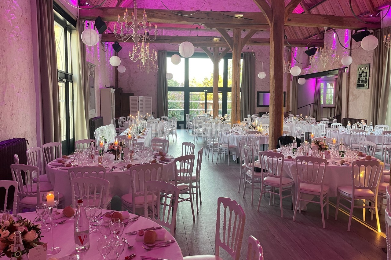 Salle de réception décorée pour un mariage, avec tables rondes dressées, chaises blanches et éclairage rose.