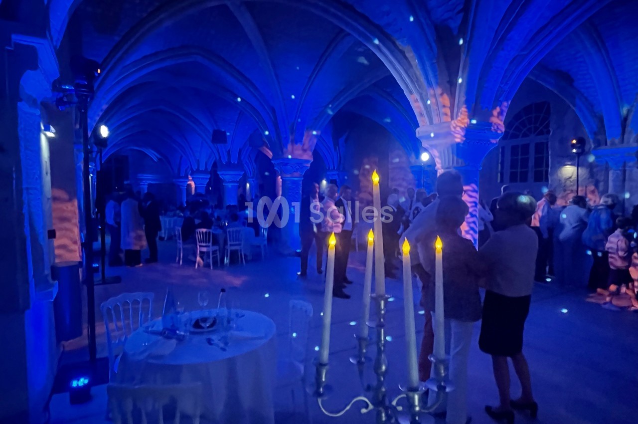 Salle voûtée éclairée en bleu avec des invités debout et des tables dressées, ambiance de réception élégante.