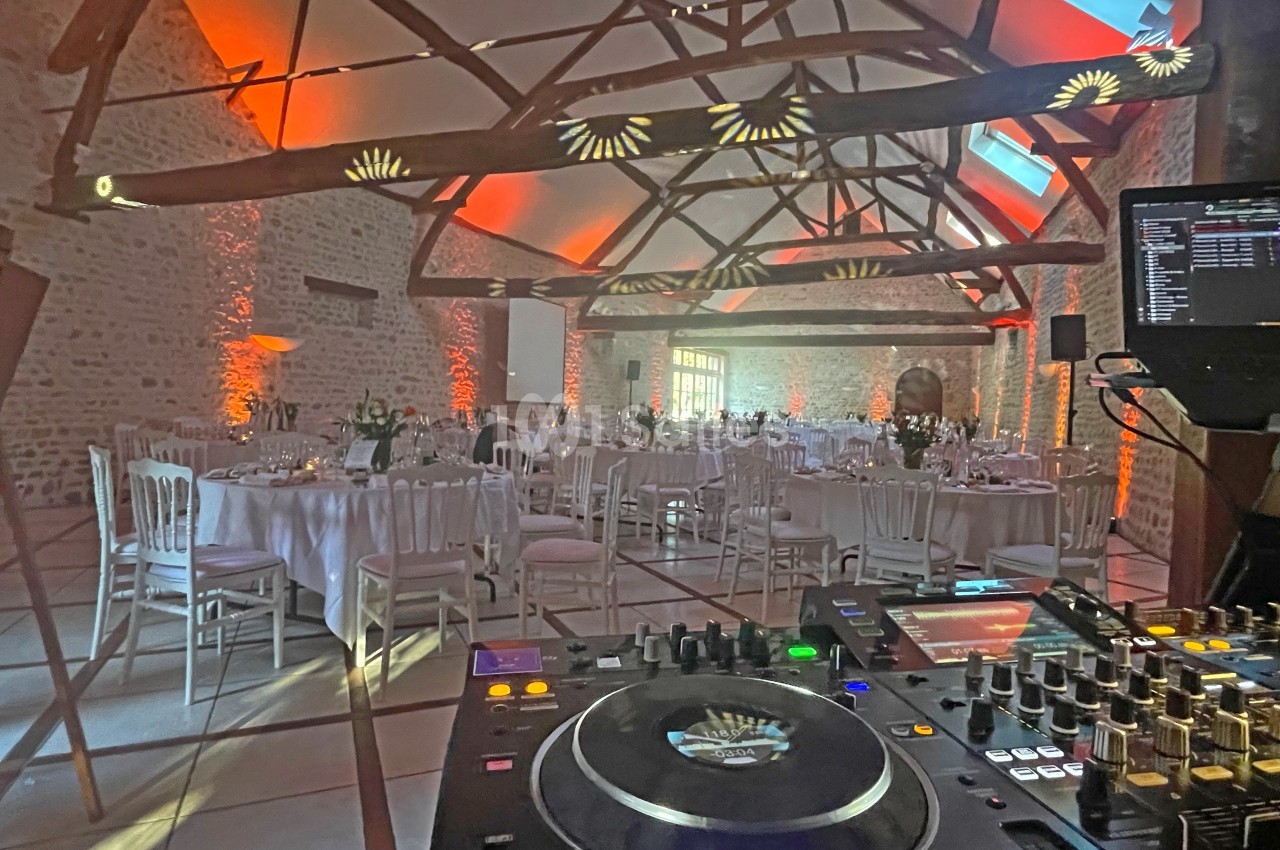 Salle de réception en pierre avec tables dressées, éclairages chaleureux et équipement de DJ au premier plan.
