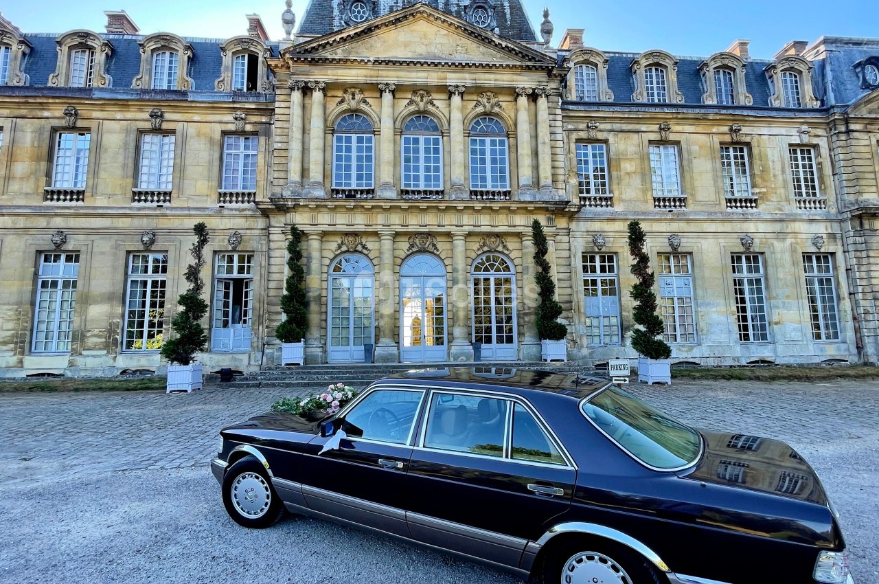 Voiture noire garée devant un château en pierre avec des fenêtres à arcades et un toit mansardé.