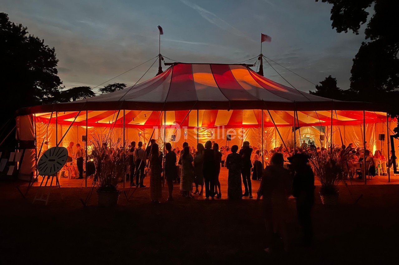 Tente de cirque éclairée en rouge au crépuscule, avec des silhouettes de personnes rassemblées à l'extérieur.