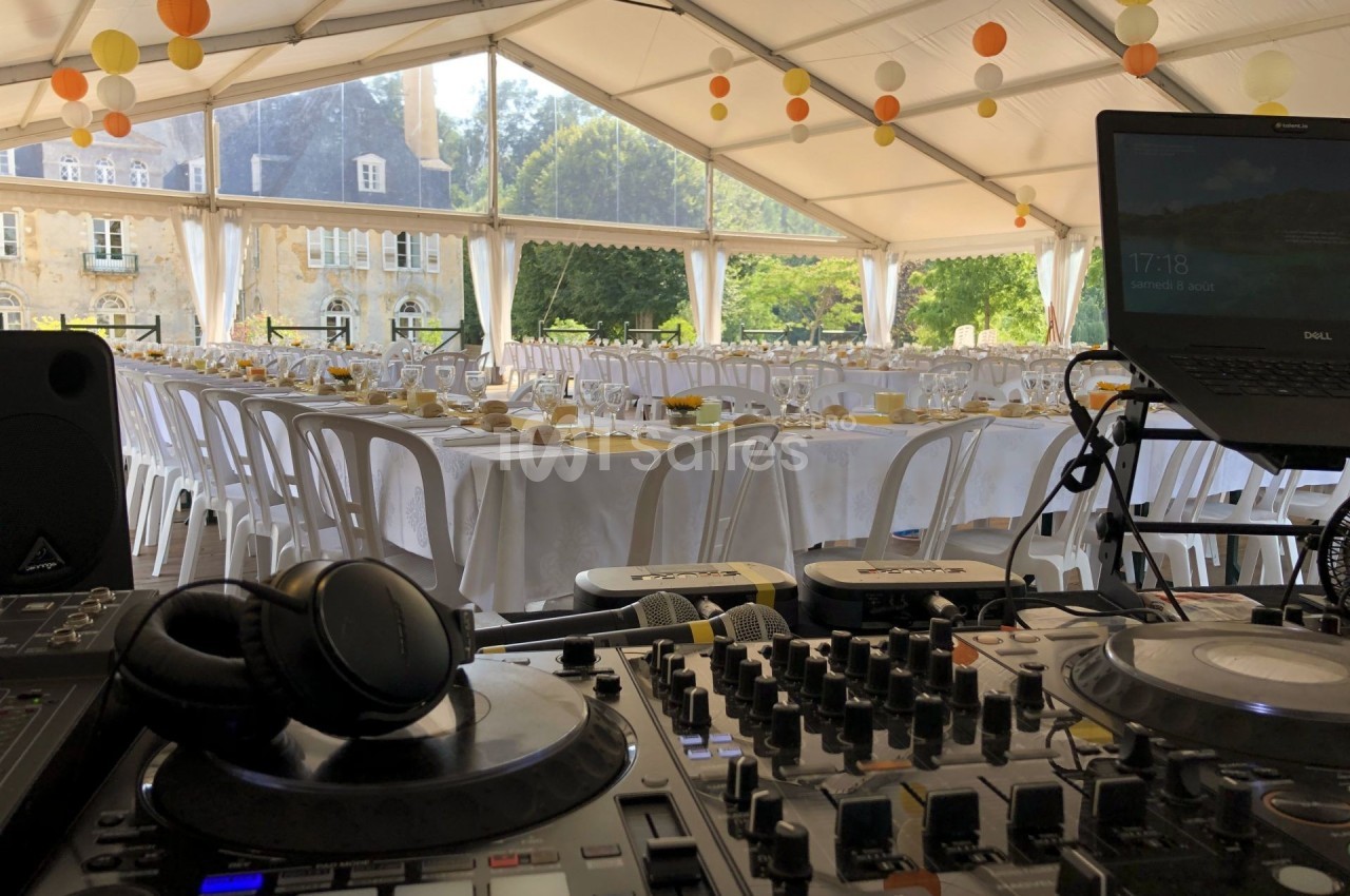 Tables dressées pour un événement sous une tente, avec une console de DJ au premier plan et un bâtiment en arrière-plan.
