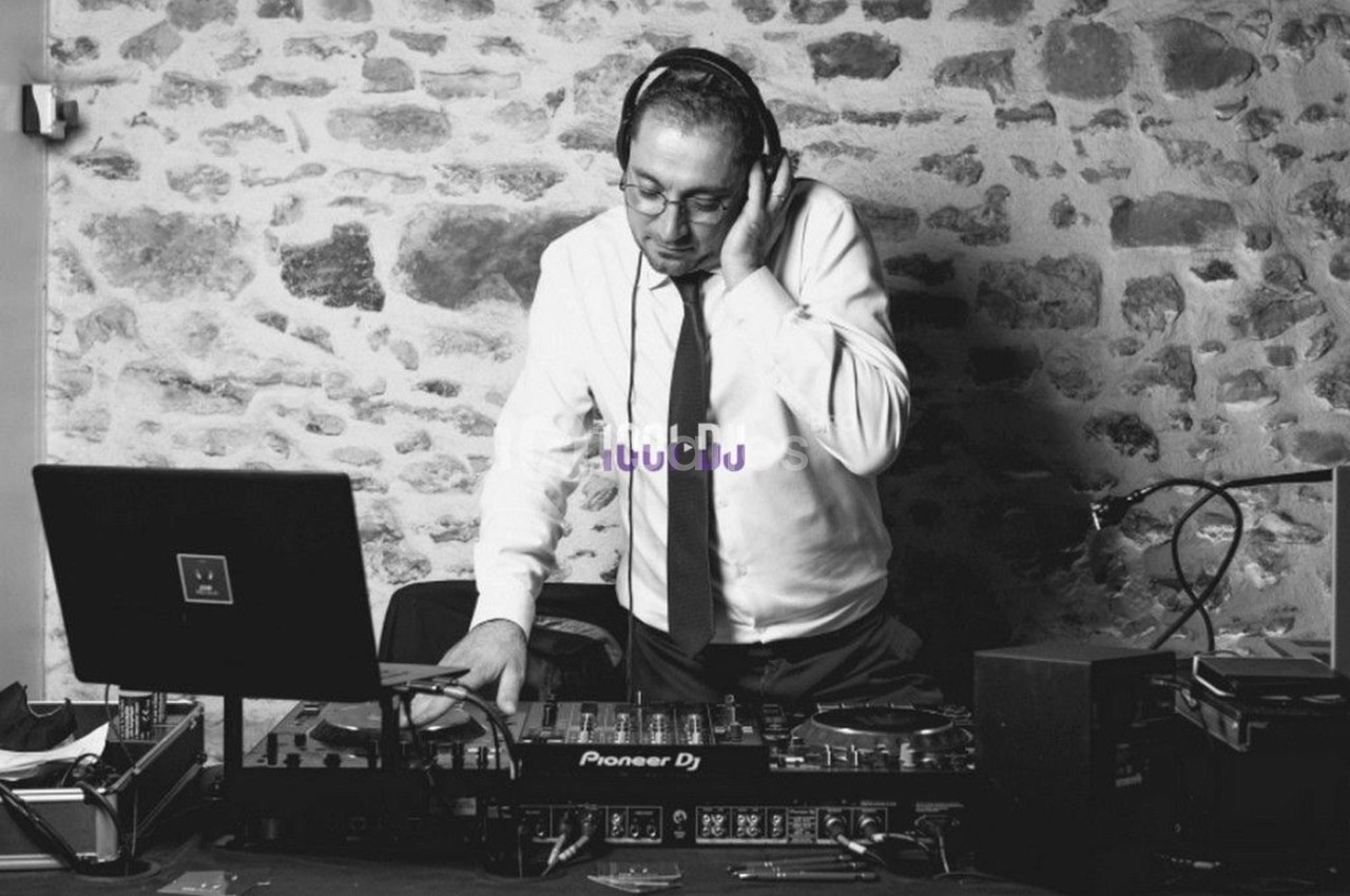 Un DJ en chemise et cravate mixe de la musique sur une table de mixage devant un mur en pierre.