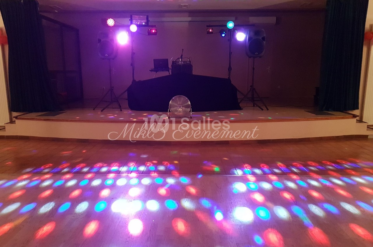 Scène de soirée avec éclairage coloré, piste de danse illuminée et équipement audio sur une estrade.