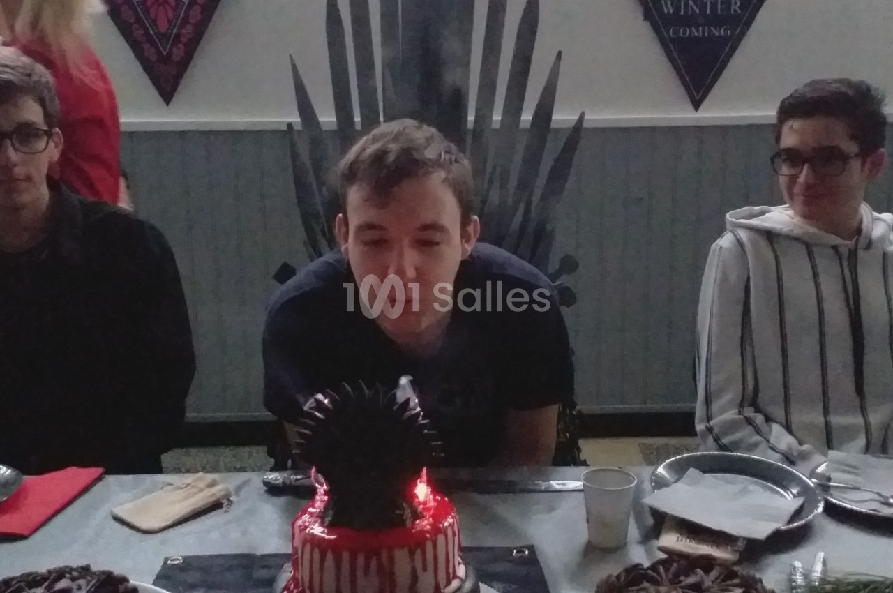 Un jeune homme souffle une bougie sur un gâteau décoré, entouré de deux personnes assises à une table.