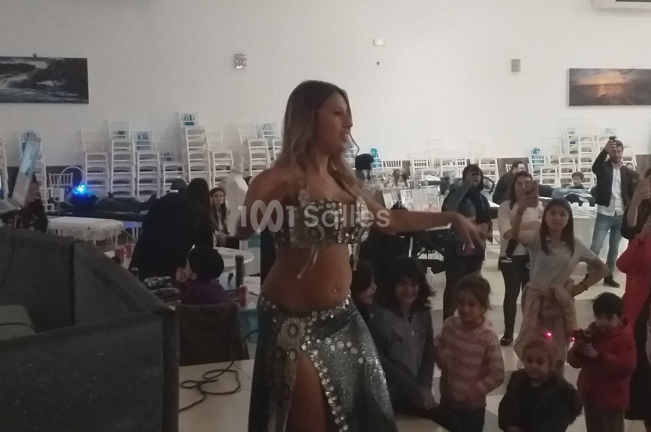 Une danseuse orientale en costume scintillant se produit devant un public composé d'enfants et d'adultes dans une salle.