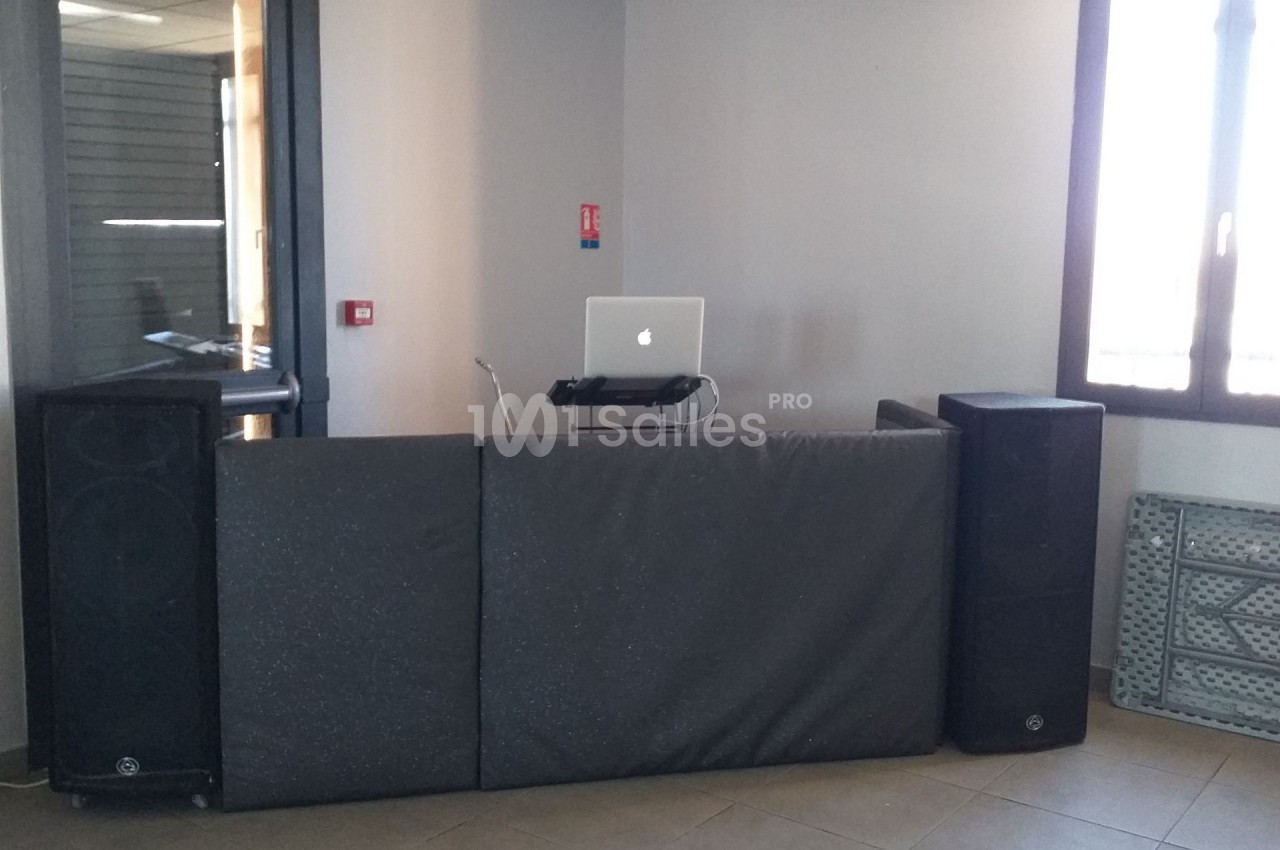 Poste de DJ avec ordinateur portable, enceintes et table recouverte de panneaux noirs dans une pièce lumineuse.