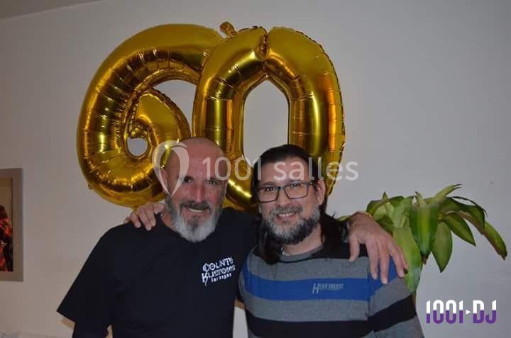Deux hommes souriants posent devant des ballons dorés formant le chiffre 60, dans une pièce intérieure.