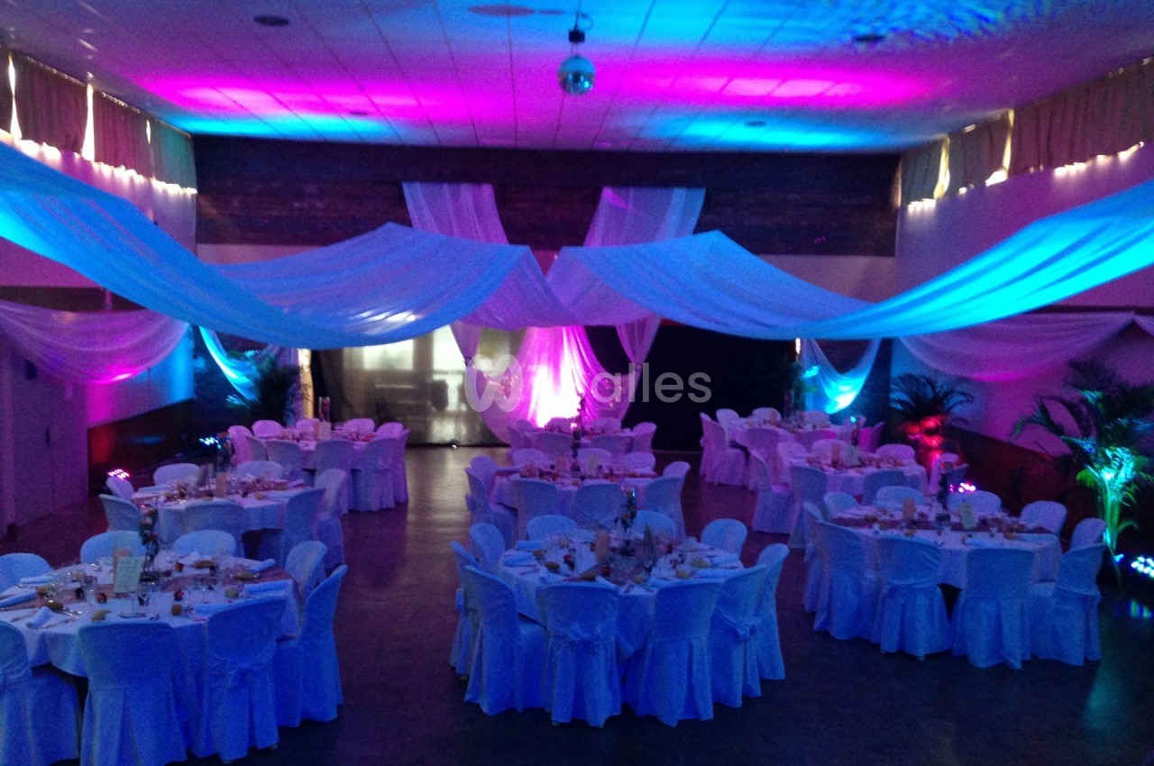 Salle de réception décorée avec des tables rondes, nappes blanches et éclairages colorés créant une ambiance festive.