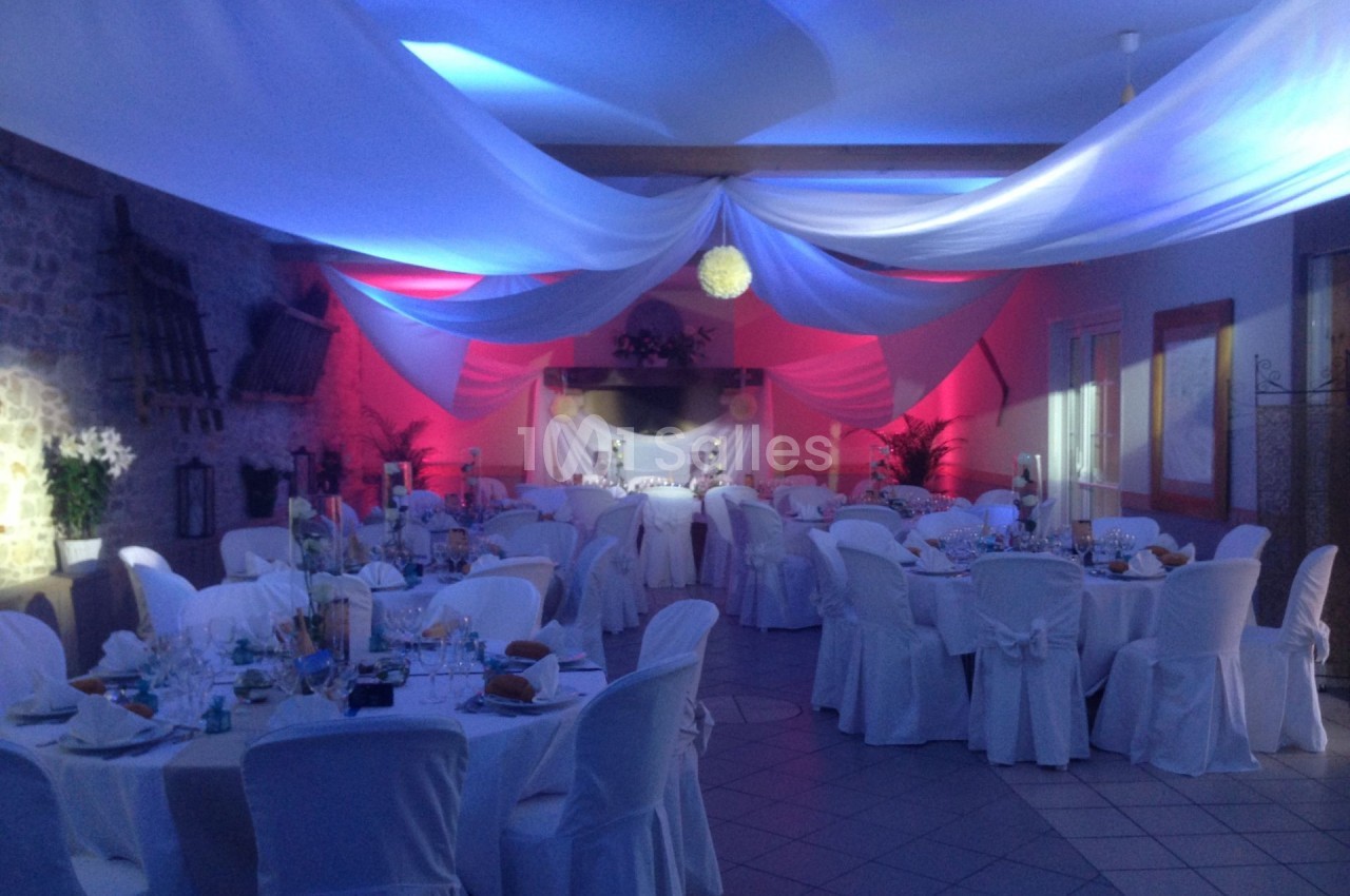 Salle de réception décorée avec des nappes blanches, des éclairages colorés et des drapés suspendus au plafond.