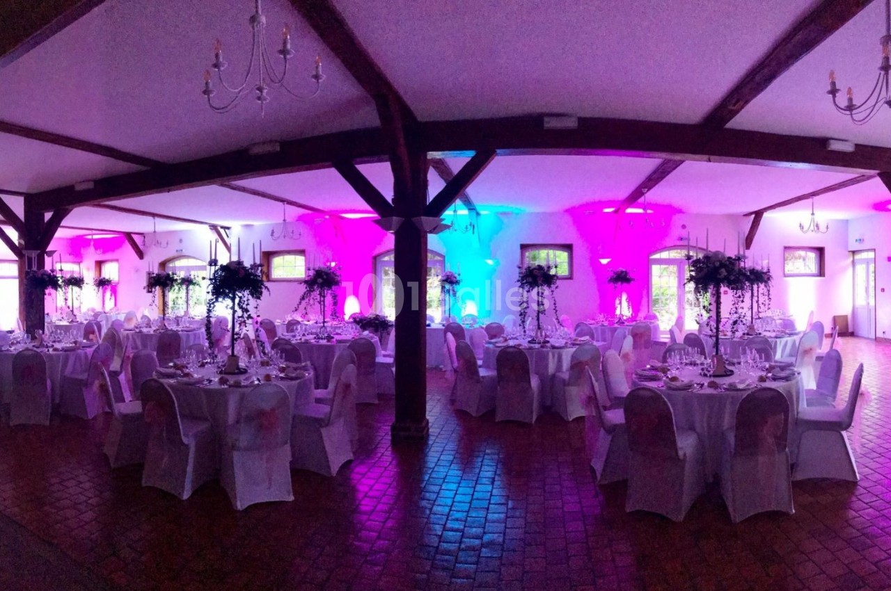 Salle de réception décorée avec des tables rondes, nappes blanches, centres de table fleuris et éclairage coloré.