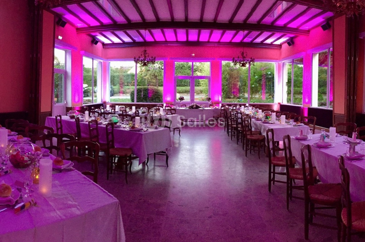 Salle de réception décorée avec des tables dressées, éclairage rose et grandes fenêtres donnant sur un jardin.