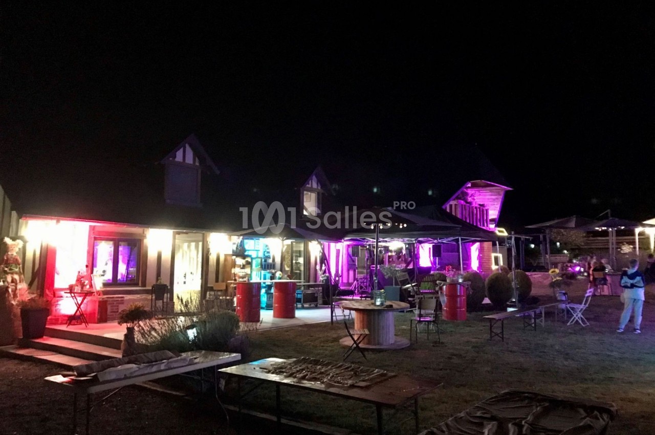 Bâtiment éclairé par des lumières colorées la nuit, avec des tables et parasols disposés à l'extérieur.