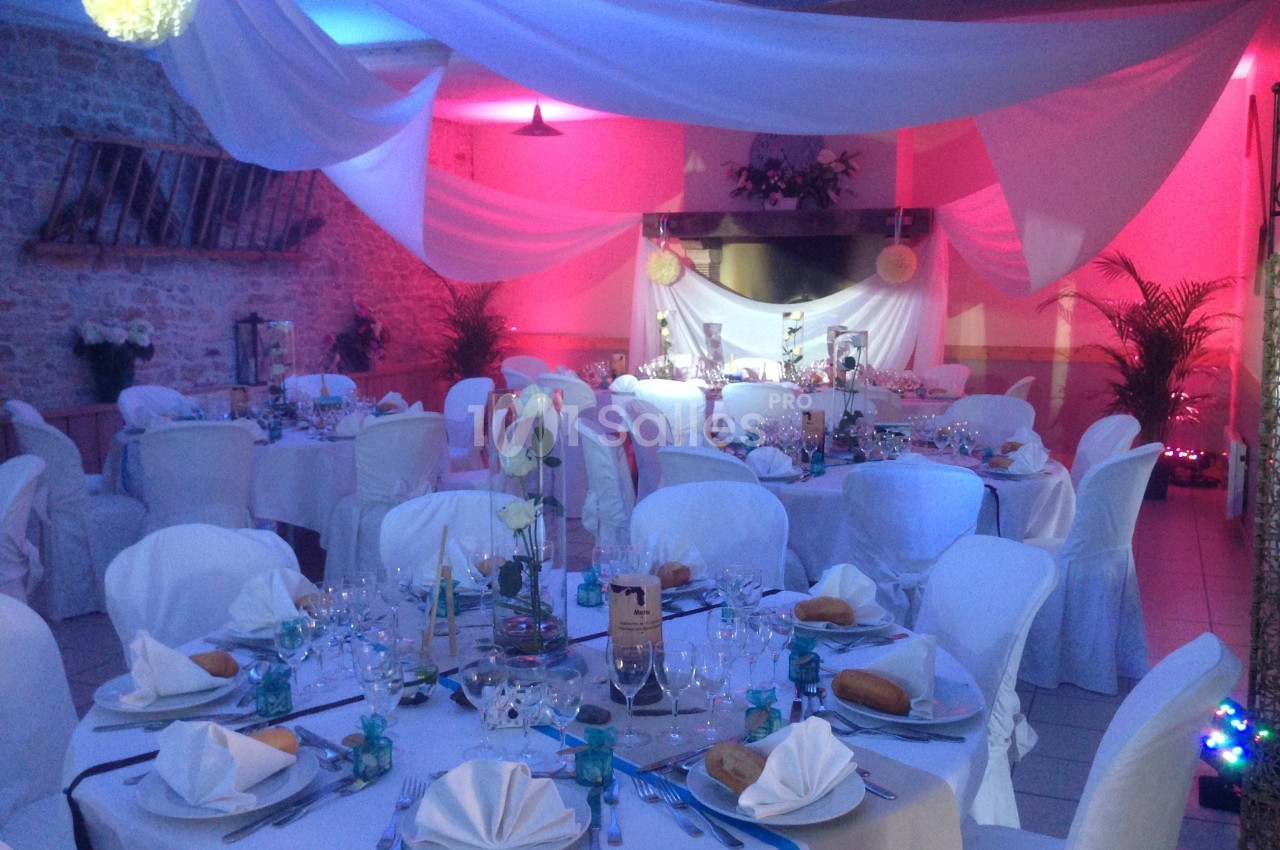 Salle de réception décorée avec des nappes blanches, des tables dressées et un éclairage rose et bleu.