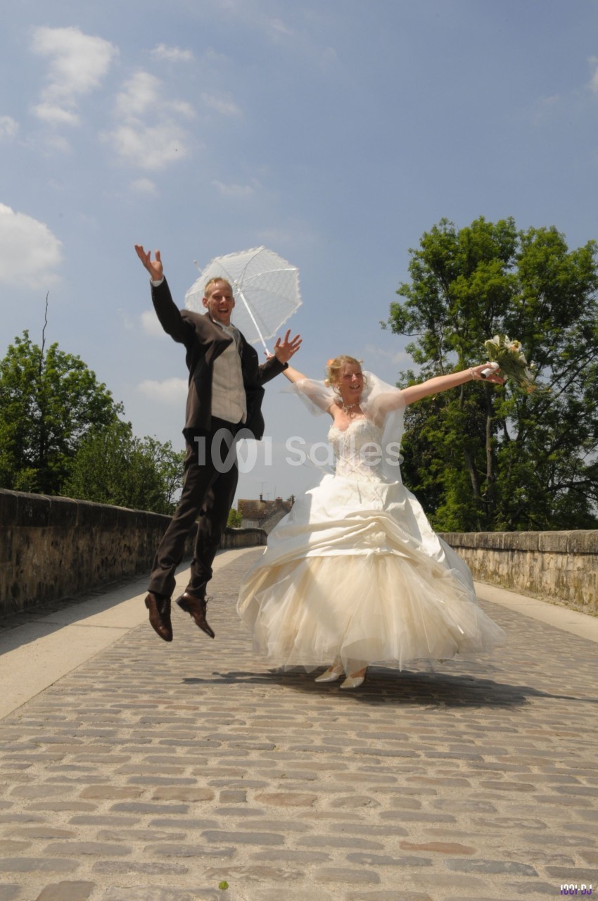 Un couple en tenue de mariage saute joyeusement sur un pont pavé par une journée ensoleillée.