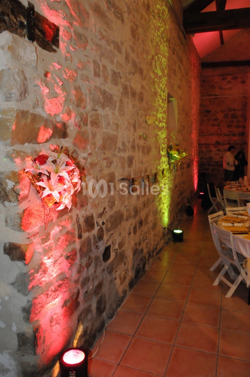 Mur en pierre éclairé par des lumières colorées, décoré de fleurs et de photos, dans une salle avec des tables dressées.