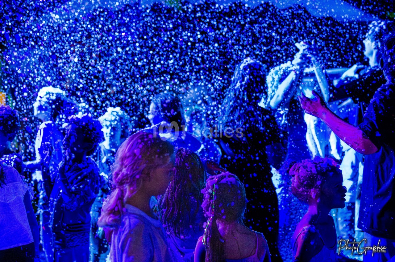 Des enfants et adultes jouent sous une pluie de mousse illuminée par des lumières bleues dans une ambiance festive.