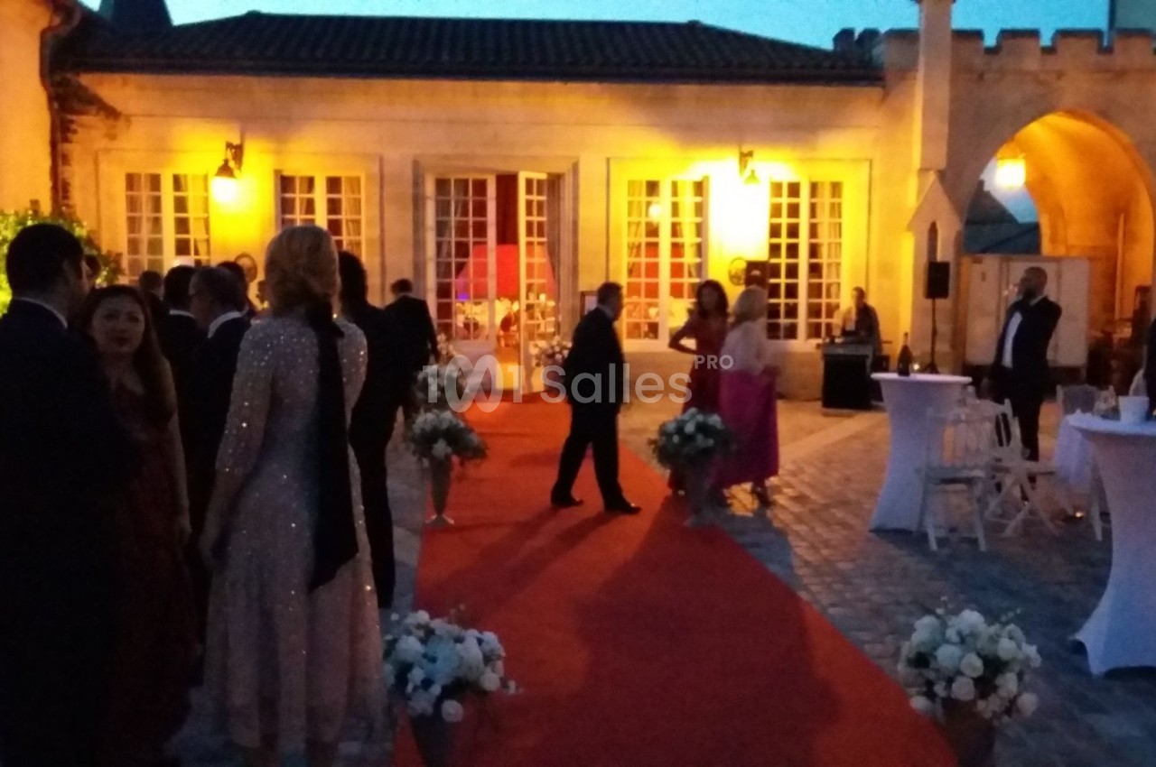 Personnes en tenue de soirée discutant sur un tapis rouge dans une cour éclairée au crépuscule.