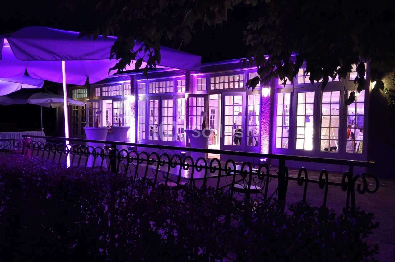 Façade d'un bâtiment éclairé en violet la nuit, avec des parasols et une clôture au premier plan.