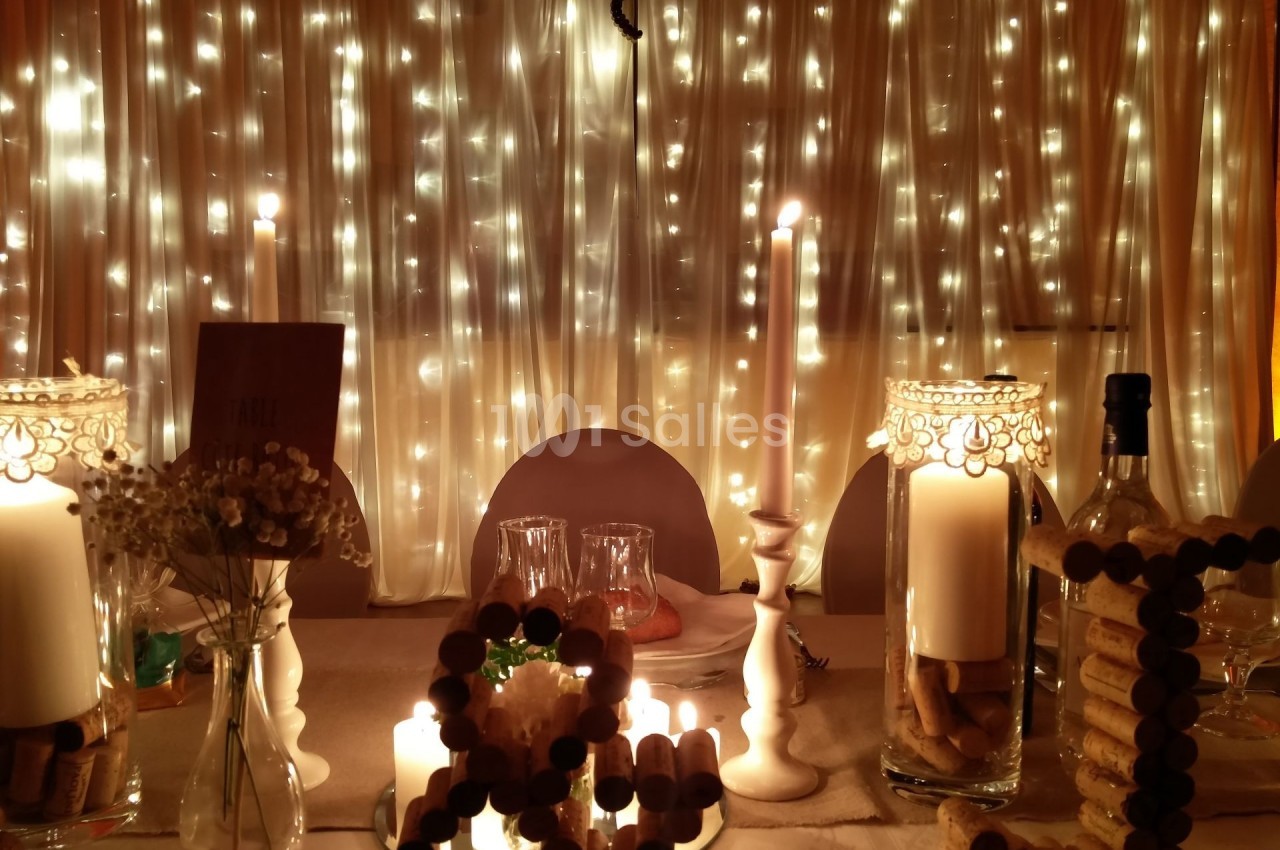 Table décorée avec des bougies, des guirlandes lumineuses et des éléments en liège, devant un rideau éclairé.