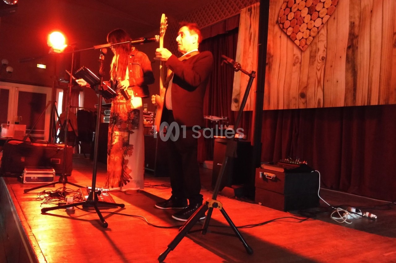 Un duo de musiciens joue sur une scène éclairée en rouge, avec un décor en bois et un cœur en rondins en arrière-plan.