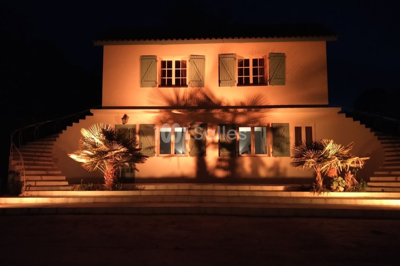 Façade d'une maison éclairée de nuit, avec des palmiers projetant des ombres sur les murs.
