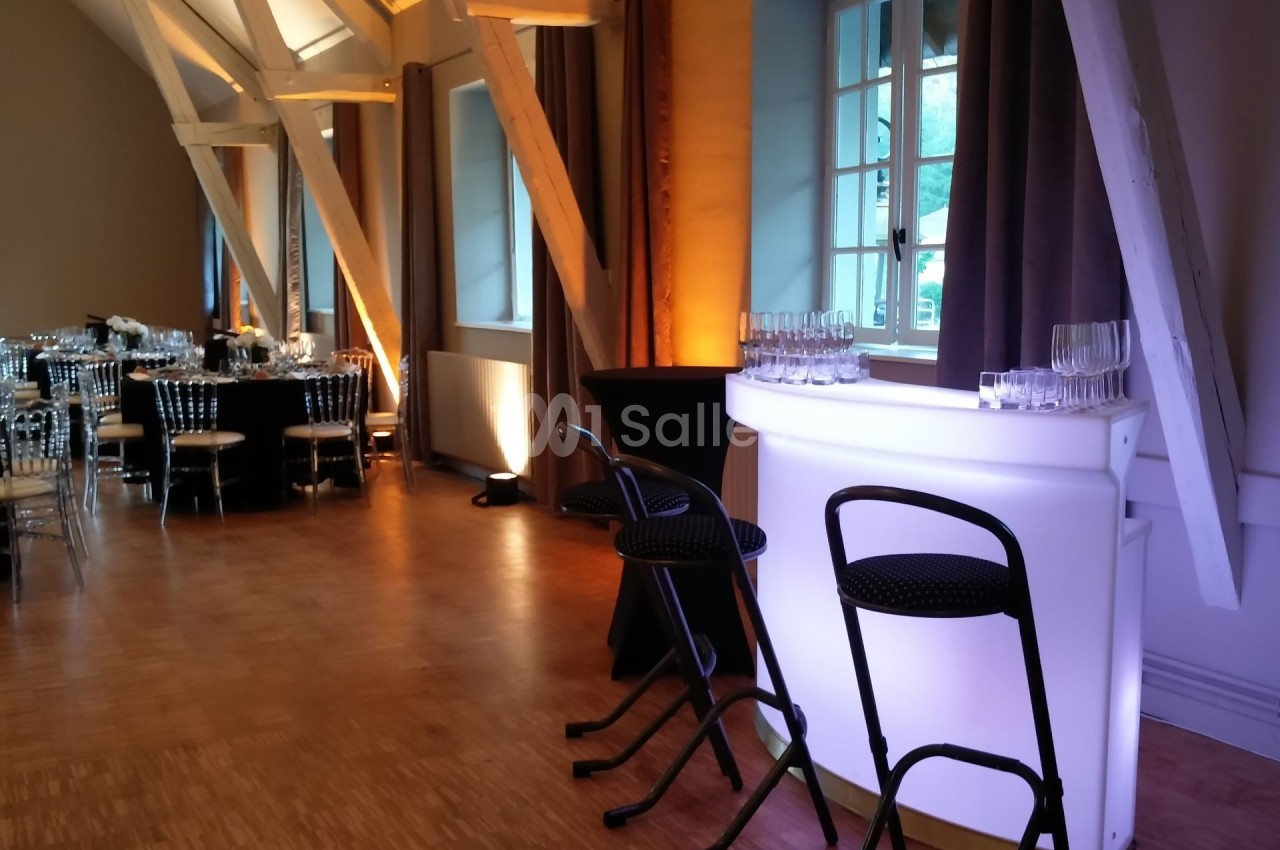Salle de réception avec tables dressées, bar lumineux blanc et tabourets noirs près de grandes fenêtres.