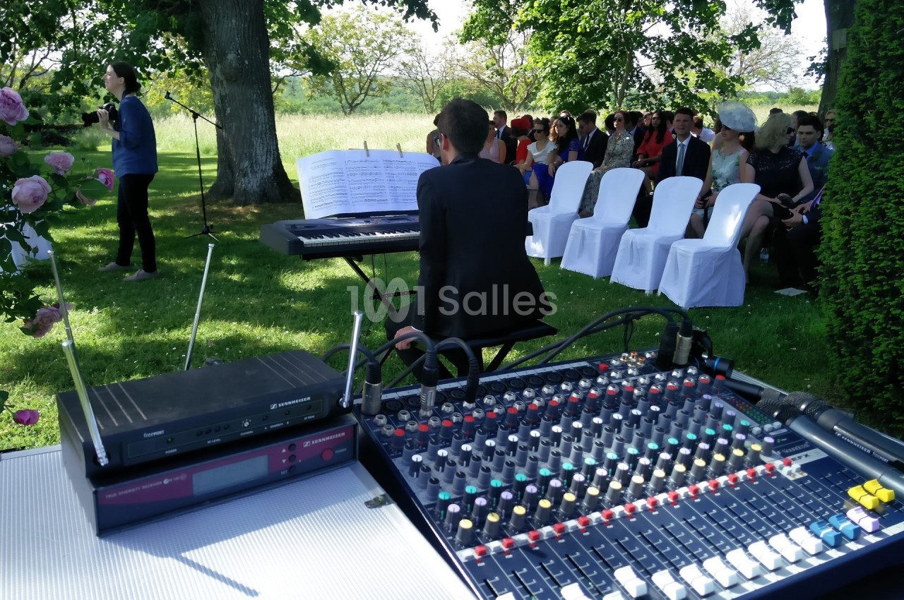 Console de mixage audio en premier plan, pianiste jouant devant un public assis en extérieur sous des arbres.