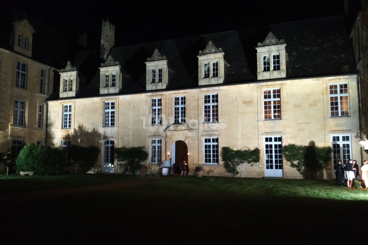 Façade d'un bâtiment historique en pierre éclairée la nuit, entourée de végétation et d'une pelouse.