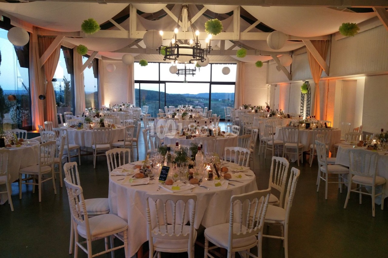 Salle de réception décorée pour un événement, avec tables rondes dressées, chandeliers et vue sur l'extérieur.