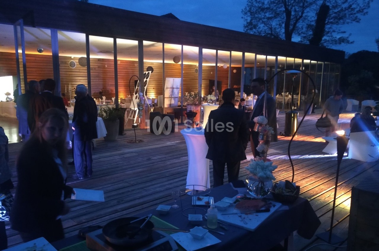 Personnes rassemblées sur une terrasse en soirée, devant une salle vitrée illuminée, ambiance conviviale.