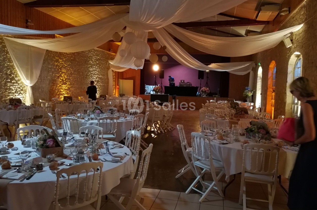 Salle de réception décorée avec des tables rondes dressées, des chaises blanches et des drapés suspendus au plafond.