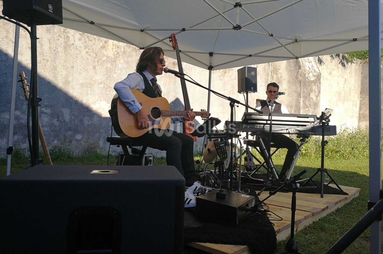Deux musiciens jouent sous une tente blanche, l'un à la guitare acoustique, l'autre au clavier, en extérieur.