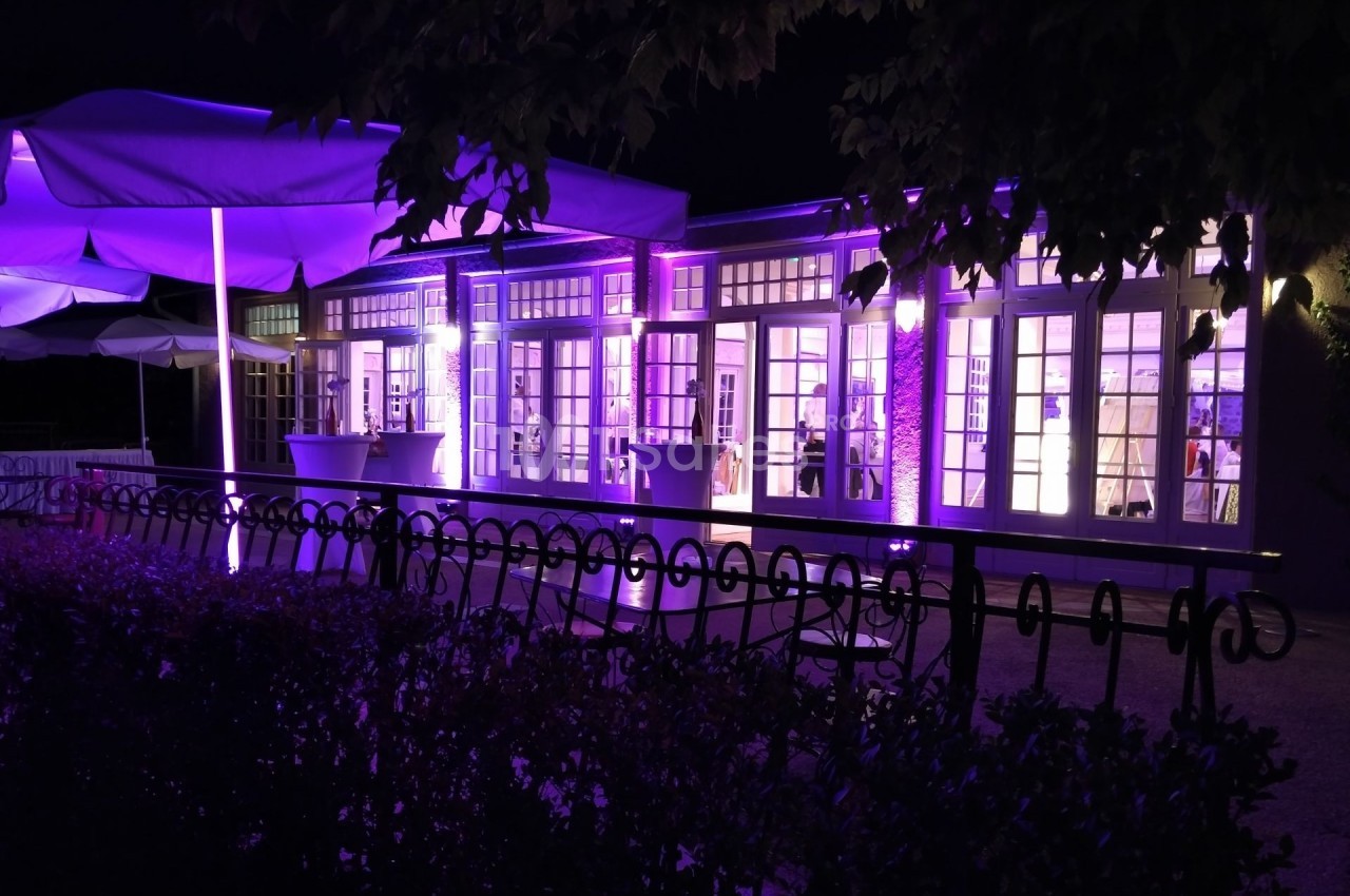 Façade d'un bâtiment éclairé en violet la nuit, avec des parasols et une clôture au premier plan.
