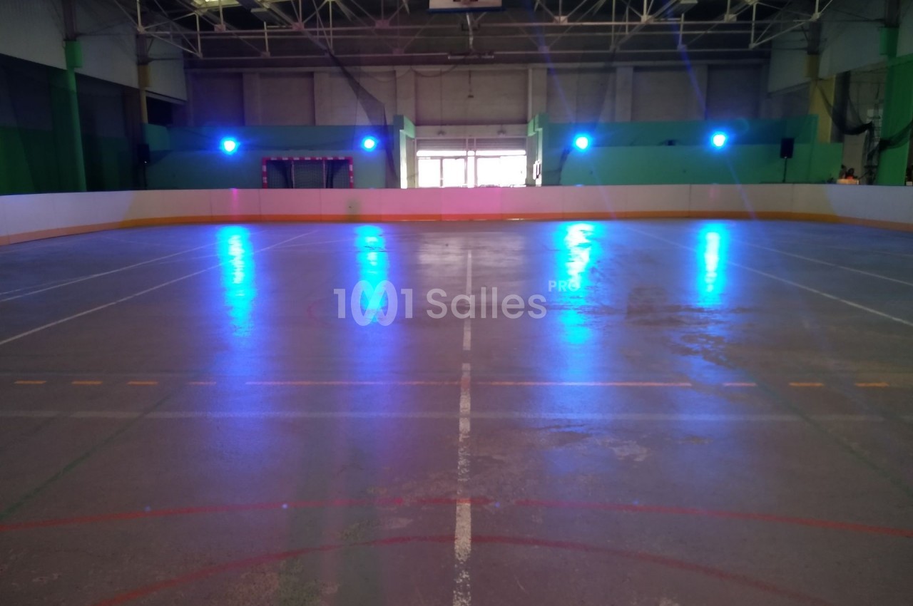 Patinoire intérieure vide avec éclairage bleu, lignes marquées au sol et structure métallique visible au plafond.